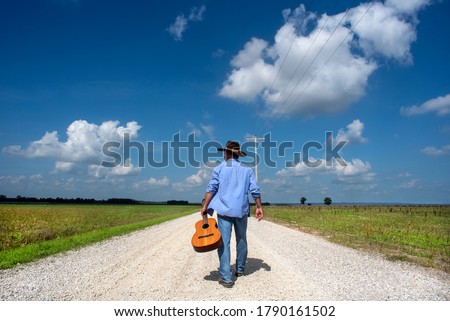 Man wearing cowboy hat walking down one lane dirt gravel country road alone, acoustic guitar, blue sky, white clouds, independent, traveling, music, rural country, back to camera, portrait