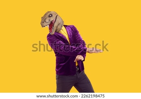 Man wearing dinosaur head mask dancing on yellow background. Stylish energetic man wearing stylish colored clothes and t-Rex animal head mask standing with arms crossed in front of him