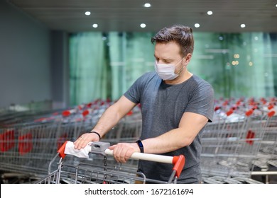 Man Wearing Disposable Medical Face Mask Wipes The Shopping Cart Handle With A Disinfecting Cloth In Supermarket. Safety During Coronavirus Outbreak. Epidemic Of Virus Covid.