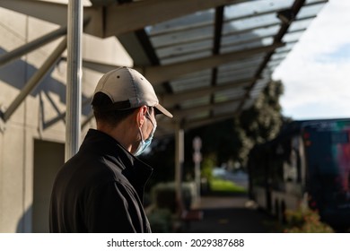 Man wearing a face mask waiting at bus stop shelter with an out-of-focus  bus stopped at roadside 