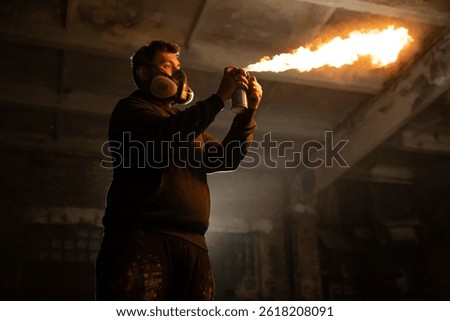 A man wearing a gas mask stands in an abandoned building, directing a spray can that emits a bright flame. The dim environment highlights the intensity of the fire.
