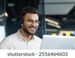 A man wearing glasses and a headset smiles while working on his laptop, embodying a cheerful and dedicated office professional in a modern work setting.