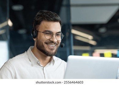 A man wearing glasses and a headset smiles while working on his laptop, embodying a cheerful and dedicated office professional in a modern work setting. - Powered by Shutterstock