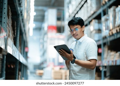 A man wearing glasses is looking at a tablet in a warehouse. He is focused on the screen and he is working