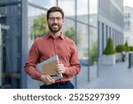 A man wearing glasses and a red shirt is holding a laptop and a piece of paper. He is smiling and standing in front of a building