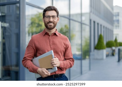 A man wearing glasses and a red shirt is holding a laptop and a piece of paper. He is smiling and standing in front of a building - Powered by Shutterstock