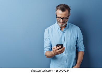Man wearing glasses standing smiling as he reads a text message on his mobile phone against a blue studio background with copy space