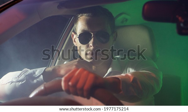A man\
wearing glasses and a white shirt sits behind the wheel of a car.\
Smoke in the cabin, multicolored light, night, a look from under\
the glasses, a portrait of the\
driver.