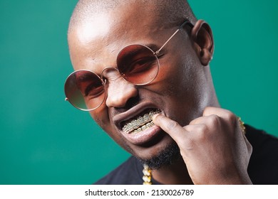 Man Wearing Gold Teeth Jewellery, Also Known As Teeth Grillsteeth Slugs, Photographed Against A Dark Green Background.