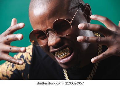 Man Wearing Gold Teeth Jewellery, Also Known As Teeth Grillsteeth Slugs, Photographed Against A Dark Green Background.