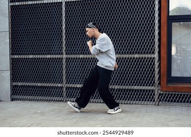 A man wearing a gray sweater and black pants dances in front of a chain-link fence in the daytime - Powered by Shutterstock