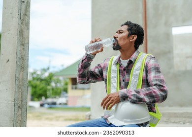 Man wearing green reflective vest Take off your helmet. and drink water to cool down After working on a construction site. An Indian foreman with a mustache sits and relaxes. - Powered by Shutterstock