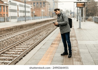 A man wearing a grey jacket stands on a train station platform, checking his phone while waiting for a train. - Powered by Shutterstock