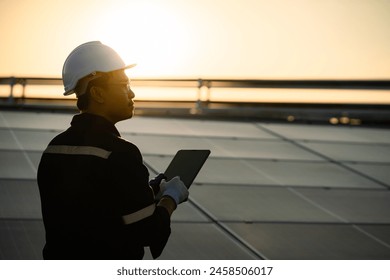 A man wearing a hard hat and safety glasses is holding a tablet in his hand. He is looking up at the sky, possibly at the sun. Concept: Renewable energy, clean technology, electric energy - Powered by Shutterstock