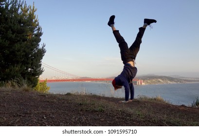 Man wearing hat, hoodie, long pants and shoes Handstands in on hill in front of the Golden Gate Bridge on the Marin side with San Francisco, California in the distance.