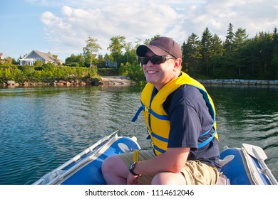 A Man Wearing A Hat And A Lifejacket Grins With Glee As He Drives A Small Blue Dinghy Around A Lake Or Sea. 