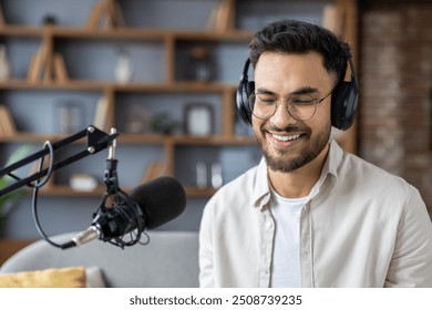 Man wearing headphones smiling while recording podcast in studio. Focused on microphone and audio production, showcasing passion for broadcasting and connection with audience. - Powered by Shutterstock