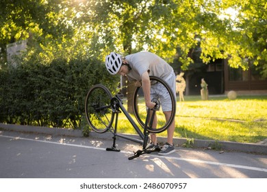 man wearing a helmet fixes a flat tire on a bicycle while standing on a paved road in a park - Powered by Shutterstock