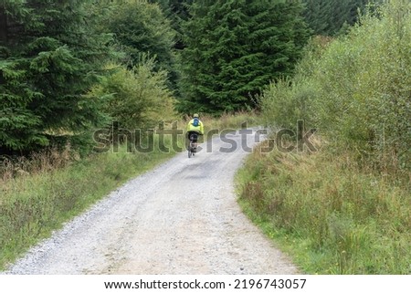 Man wearing a high viz yellow vest riding a mountain bike through the forest trail. Grey gravel path with lush green trees at the sides.
