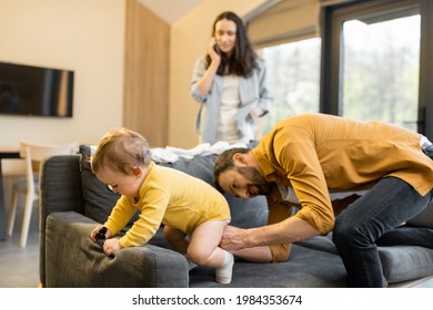 Man Wearing His Baby Son On The Couch At Home. Fatherhood And Child Care By Father Concept. Wife Talks On Phone On The Background