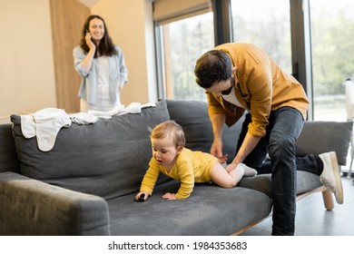 Man Wearing His Baby Son On The Couch At Home. Fatherhood And Child Care By Father Concept. Wife Talks On Phone On The Background