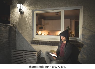 A Man Wearing A Hoodie Sits Outside The Kitchen In The Garden Under An Outdoor Security Light As He Adjusts The Light Output From His Digital Tablet At Night Time In A House In Edinburgh, Scotland, UK