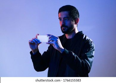 Man Wearing Jacket And Holding Cell Phone Illuminated By Blue And Red Lights. Concept Of Man Filming The Police On His Smartphone