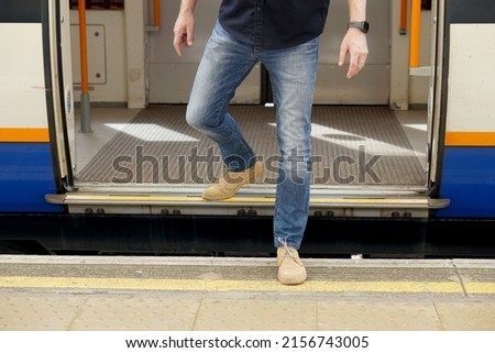 Man wearing jeans, blue shirt and light brown shoes stepping down from the train to platform at the train station