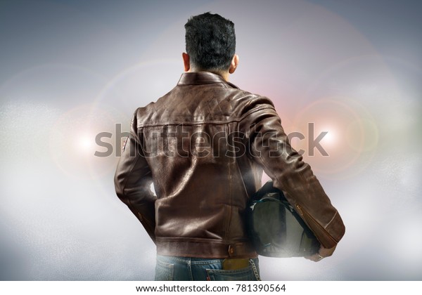a man wearing a\
leather jacket and holding a motorcycle helmet standing in front of\
the car light.
