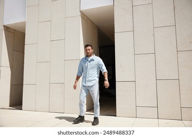 A man wearing a light blue shirt and jeans walks out of a modern building, looking confident and focused. - Powered by Shutterstock