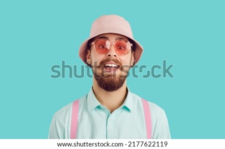 Man wearing pink bucket hat, cool stylish sunglasses, shirt and suspenders isolated on turquoise background looking at camera with funny happy surprised amazed face expression. Summer fashion concept