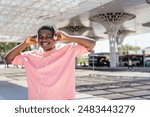 A man wearing a pink shirt is listening to music with headphones. He is smiling and he is enjoying himself. The scene takes place in an urban setting, with a bus.