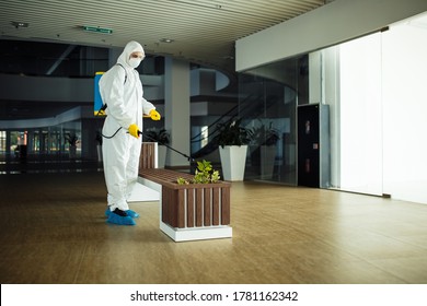 A Man Wearing Protective Suit Is Disinfecting A Bench In An Empty Shopping Mall With Sanitizing Spray. Cleaning Up The Public Place To Prevent Covid Spread. Healthcare Precautions And Safety Concept.