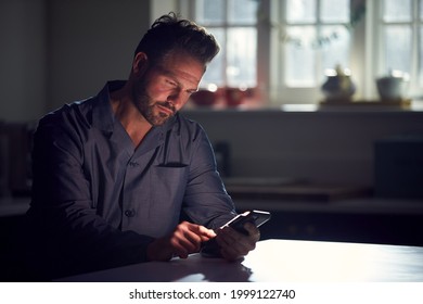 Man Wearing Pyjamas Sitting In Kitchen At Night Using Mobile Phone