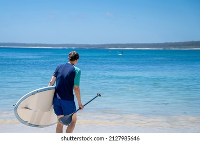 Man Wearing Rash Guard With Stand-up Paddle Board On A Sandy Beach Near The Ocean Bay In Australia. SUP Water Sport Summer Activity