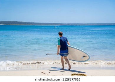 Man Wearing Rash Guard With Stand-up Paddle Board On A Sandy Beach Near The Ocean Bay In Australia. SUP Water Sport Summer Activity