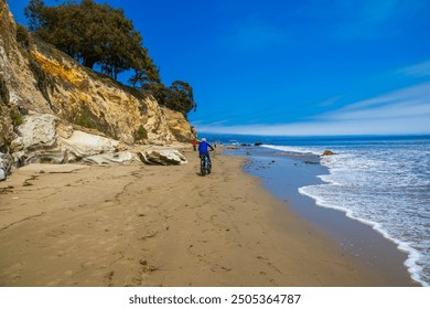 a man wearing a red jacket riding an electric bike on the brown sandy beach with people walking along the beach, blue ocean water and lush green trees at Leadbetter Beach in Santa Barbara California	 - Powered by Shutterstock