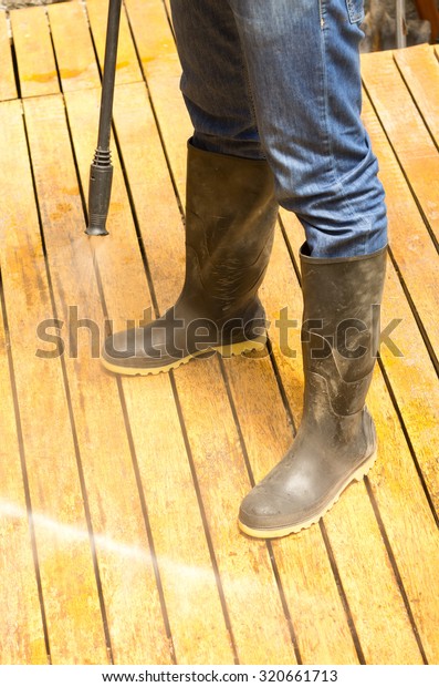 Man wearing rubber boots using high\
water pressure cleaner on wooden terrace\
surface.