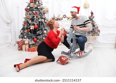 A man wearing a Santa hat sits on a rocking horse while toasting with a woman in a red sweater. They are celebrating Christmas surrounded by presents and a decorated Christmas tree - Powered by Shutterstock