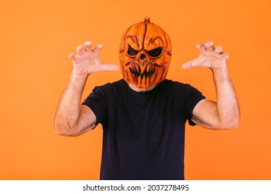 Man wearing scary pumpkin latex mask with blue t-shirt scares with his hands, on orange background. Halloween and days of the dead concept.