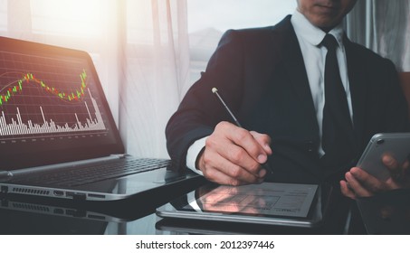 A Man Wearing A Suit On His Left Hand Holding A Cell Phone His Right Hand Poked A Pen On A Tablet's Screen, And The Screen Of The Laptop Shows The Fast-growing Stock Chart.