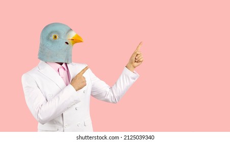 Man wearing trendy white suit and funny pigeon bird mask standing isolated on solid pastel pink colour background and pointing both index fingers at blank marketing text copyspace on right side