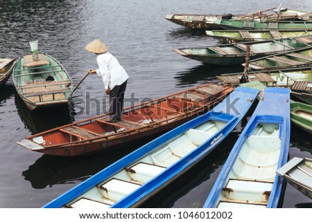 Man wearing white shirt, conical hat with his paddle stand on red empty rowing boat with many boats stop over the river at Trang An Grottoes in Ninh Binh, Vietnam.