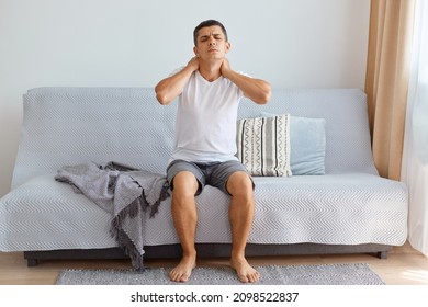 man wearing white T-shirt sitting on sofa in living room, touching neck feeling pain and numbness, worried about muscle tension, osteochondrosis, uncomfortable sleeping conditions.