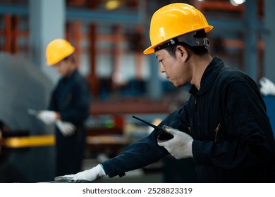 A man wearing a yellow hard hat and a black jacket is holding a remote control. He is standing in front of a large metal structure - Powered by Shutterstock