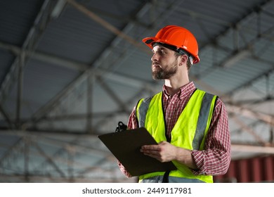 A man wearing a yellow vest and an orange hard hat is holding a clipboard. He is looking up at something, possibly a ceiling or a structure. Concept of caution and attention to detail - Powered by Shutterstock