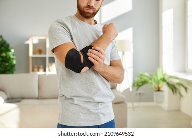 Man Wears Bandage On His Elbow Which Serves As Fixator To Restore And Relieve Joint Pain. Cropped Image Of Man Kneading His Sick Elbow And Injured Arm While Standing At Home. Pain Relief Concept.