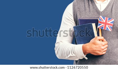 Man wears grey sweater vest holds english books and flag before dark blue studio background, language learning concept