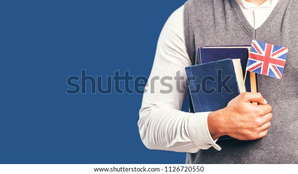 Man\
wears grey sweater vest holds english books and flag before dark\
blue studio background, language learning\
concept