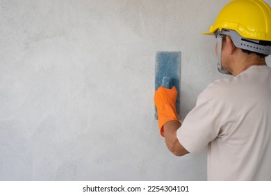 A man wears hard hat and gloves holding trowel is plastering cement wall with copy space for text, concept of wall plastering, structural work, plaster work, home building, construction work 